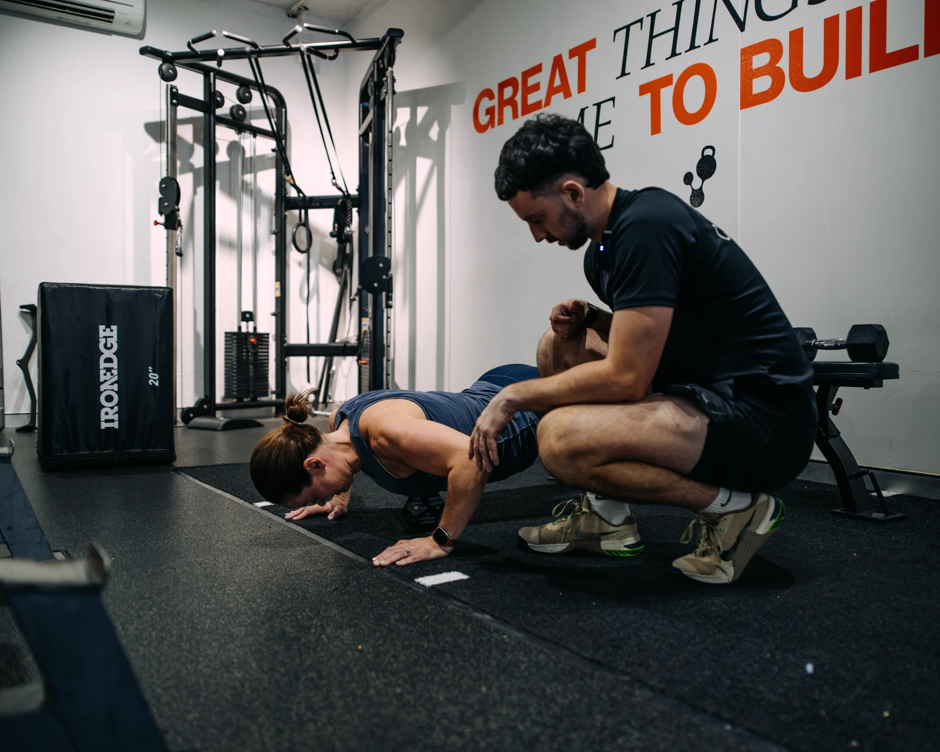 A coach crouches beside a client to guide a floor exercise.